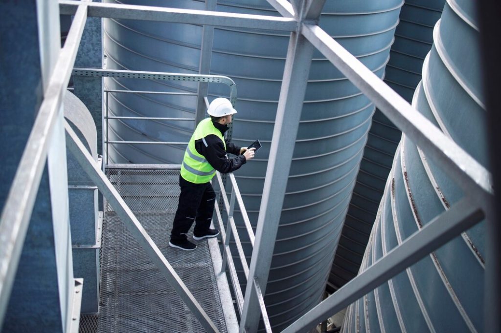 Industrial worker on metal platform inspecting storage tanks with tablet